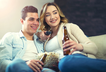 Happy young couple sitting on the sofa at home with popcorn and beer  watching TV. They are laughing and watching a movie or television.