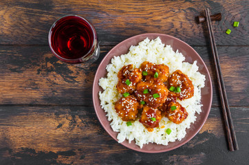 Crispy sesame chicken, chopped breast fillets, with a sticky sweet Asian sauce and white boiled rice on a plate on wooden table, top view.