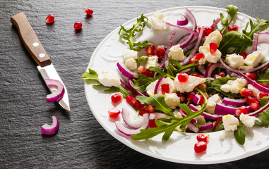 Dietary salad with red onion, arugula, feta and pomegranate seeds in white plate. Black stone background.