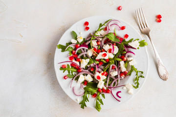 Dietary salad with red onion, arugula, feta and pomegranate seeds in white plate. Light table, top view.