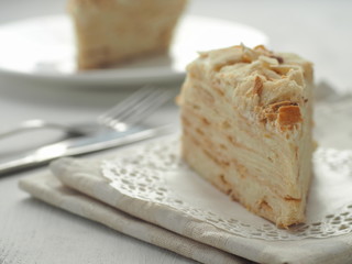 Piece of multi layered cake close-up. Mille feuille dessert. Crumbs decorated torte on white doily upon wooden table. Food background, blur. Selective focus on the front.