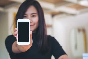 Mockup image of a beautiful woman holding and showing white mobile phone with blank black screen and smiley face in vintage cafe