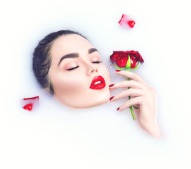 Beautiful model girl with bright makeup holding red rose flower in her hand and relaxing in milk bath
