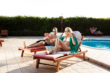 Friends lying on chaises near swimming pool, making selfie, smiling.