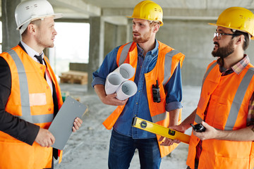 Group of three workmen wearing protective helmets and vests standing among concrete walls on construction site discussing development progress with inspector