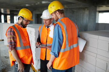 Group of three workmen wearing protective helmets and vests standing among concrete walls on...
