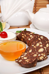 Chocolate sausage biscuit with condensed milk on a white plate on a background of a set for tea and jam
