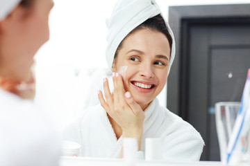 Perfect young woman applying facial cream in bathroom