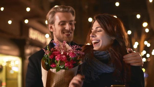 Young handsome Caucasian male woth light beard and bouquet of flowers closing eyes of beautiful female and kissing her in slowmotion