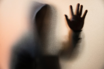 Shadow blur of horror man in jacket with hood.One hands on the glass.Dangerous man behind the frosted glass.Mystery man.Halloween background.Blur picture. - obrazy, fototapety, plakaty