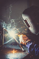 Industrial worker welding round pipe on a work table, producing smoke, sparks and colorful reflections - obrazy, fototapety, plakaty