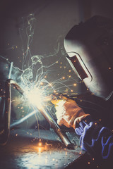 Industrial worker welding round pipe on a work table, producing smoke, sparks and colorful reflections