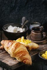 Breakfast with two croissant, butter, cottage cheese, coffee and sliced mango fruit, served on wood chopping board over old wooden background. Close up.
