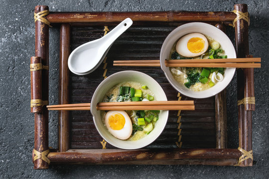 Two bowls with asian style soup with scrambled eggs, half of marinated egg, spring onion, spinach served with wood chopsticks and spoons on bamboo tray on black concrete background. Top view