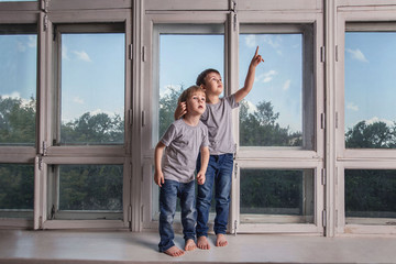 Two little boy in jeans and grey t-shirts : friends, brothers play standing on the windowsill of a huge white retro Windows. The older boy shows something to the little boy and he looks interested.