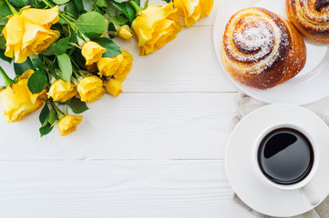 Cup of coffee with bouquet of yellow roses, flat lay, top view. Two spiral tasty buns with coconut chips, cup of black coffee and bouquet of yellow roses. Delicious breakfast, sweet rolls and coffee. 
