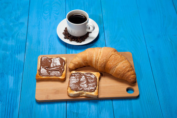 Cup of coffee with a croissant and toast with chocolate on a blue wooden table