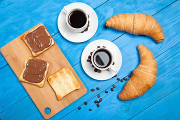 Two cups with coffee, croissant and toast with chocolate on a blue wooden table