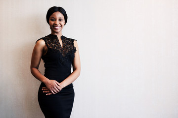 classy black woman smiling into the camera while posing in a beautiful black dress against a textured beige wall, with excitement visible in her facial expression.