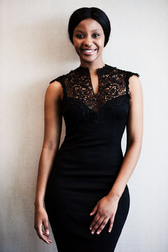 Well dressed attractive young black woman wearing a slim black dress smiling at the camera wile posing in front of a textured beige wall.