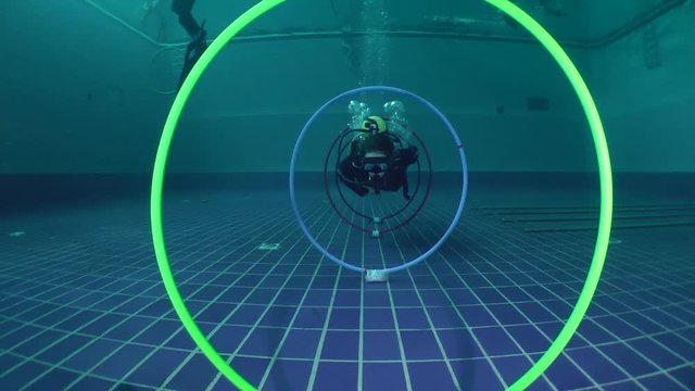 Woman diver swims in scuba gear through the rings installed on the bottom of pool.
