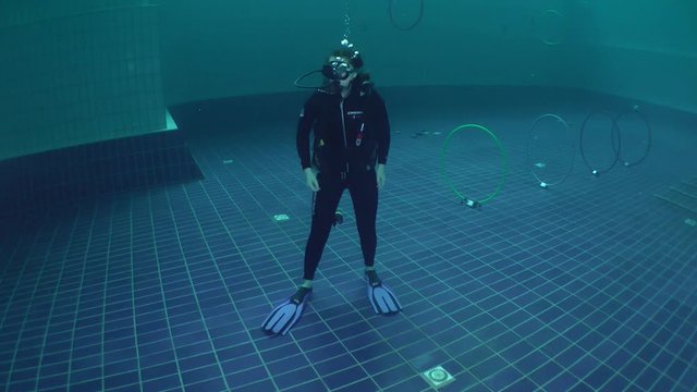 The girl-diver in scuba gear standing on the bottom of the pool, wide shot.
