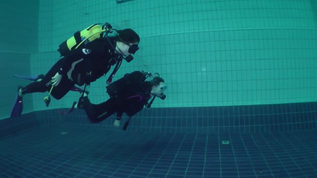 Two girls diver in scuba gear swimming along the pool wall.
