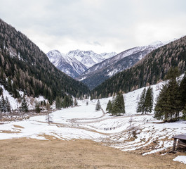 A winding road in mountain environment. The snow partially covers the ground. The trees are without snow. Mountains in the distance. Cloudy sky