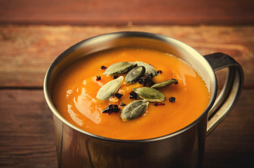 Roasted pumpkin soup in metal mug with seeds and smoked paprika, on rustic wooden background.