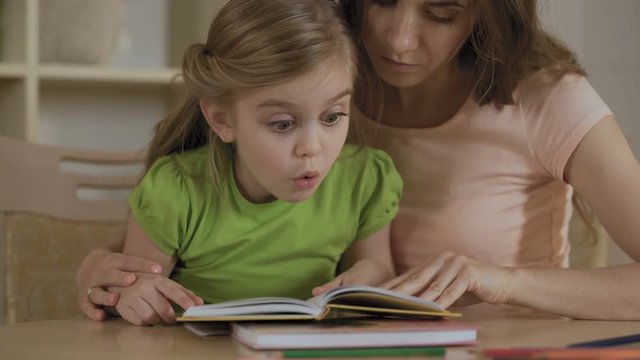 Supportive mother reading textbook together with hyperactive little daughter