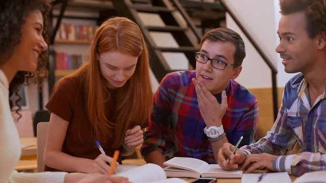 Group of four interracial friends sitting at table in library reading book making notes talking discussing in slowmotion