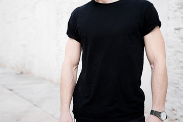 Closeup view of young muscular man wearing black tshirt and jeans posing outside. Empty white wall...