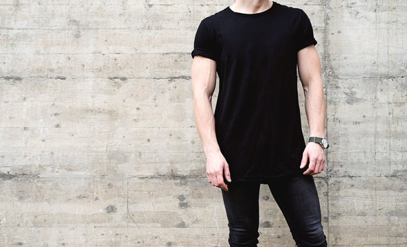 Young muscular man wearing black tshirt and jeans posing in center of modern city. Empty concrete wall on the background. Hotizontal mockup.