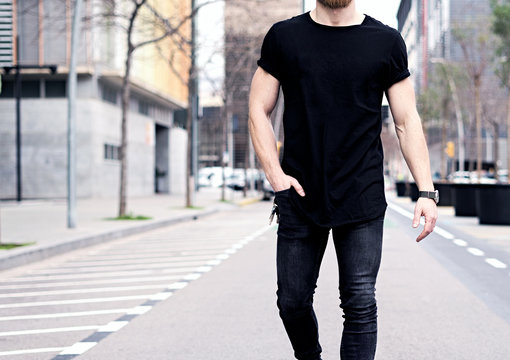 Closeup view of young muscular man wearing black tshirt and jeans walking on the streets of the modern city. Blurred background. Hotizontal mockup.