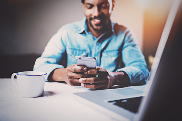 Happy African man using smartphone while sitting at the wooden table his modern home.Concept of young people working with mobile devices.Selective focus on male hands.Blurred background,flares.