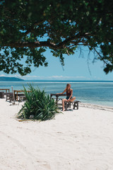 Blonde hair girl sits on the wooden chair on the beach in bikini and shorts near Indian ocean