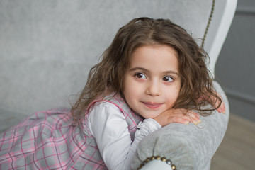 Pretty little girl with brown hair sits on a grey sofa and looks away