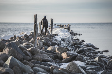 Young man standing on the rocks on the old breakwater between wooden bollards at the seaside in winter afternoon.