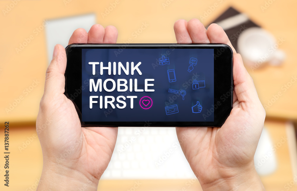 Aufkleber Watching two hand holding mobile phone with Think mobile first word and feature icon with blur desk office background,Digital marketing