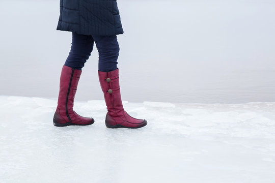 Woman walking on the seashore ice surface in winter day. Red winter boots on legs. Active lifestyle at the sea.
