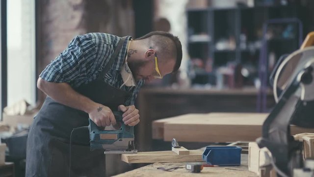 Joiner works with an electric jigsaw and processes wooden products. Stylish craftsman with brutal hairstyle and saved glasses work at his workstation. Modern woodworker, woodman with tattoo.