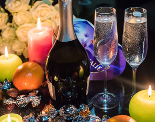 A bottle of sparkling wine or champagne, some fruits and sweets on the table with burning candles and flowers. Romantic date.