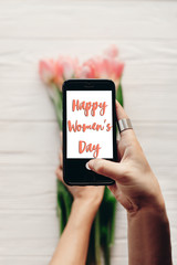 happy women's day text sign on screen of phone. hand holding gadget with message   above pink tulips on white wooden rustic background. greeting card concept