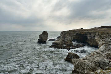 Cold winter afternoon by the sea near Tyulenovo, Bulgaria. Dark silhouettes of cliffs with caves. Waves crash into the icy cliffs.