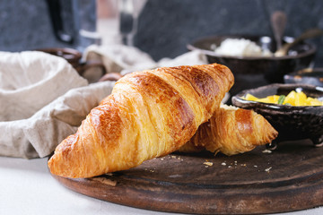 Breakfast with two croissant, butter, cup of coffee, cottage cheese and sliced mango fruit, served on serving wood board with textile napkin on white and gray concrete texture background. Close up.