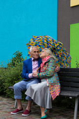 Elegantly dressed elderly couple sitting on  bench under an umbrella.