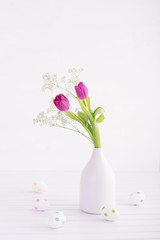 Vase of fresh tulips with decorative easter eggs on wooden table. High key