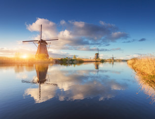 Obrazy na Szkle  Wiatraki o wschodzie słońca. Rustykalny krajobraz z niesamowitymi holenderskimi wiatrakami w pobliżu kanałów wodnych z niebieskim niebem i chmurami odbijającymi się w wodzie. Piękny poranek w Kinderdijk w Holandii na wiosnę. Podróżować