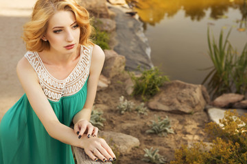 Beautiful blonde woman with curly short bob hairstyle, delicate make up and red lips in green short dress at the park. Fashion sensual posing on summer sunset. Outdoors. Copy space