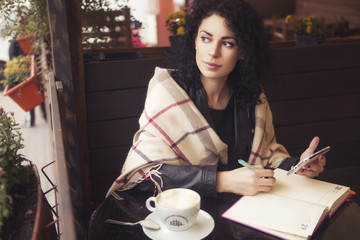 Beautifil brunette caucasian woman in leather jacket and plaid sitting on a cafe drinking coffe and working on her phone and notebook. cold weather